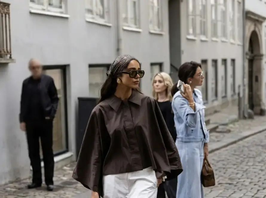 A woman in a brown wide-sleeve shirt, white trousers, and a patterned headscarf walking on a cobblestone European street during a fashion event.