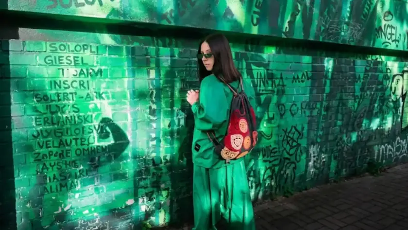 A woman in an oversized bright green streetwear outfit, sunglasses, and a colorful crochet backpack stands centered against a graffiti-covered green brick wall.