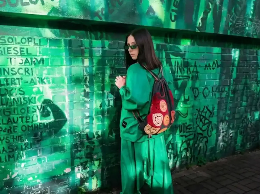 A woman in an oversized bright green streetwear outfit, sunglasses, and a colorful crochet backpack stands centered against a graffiti-covered green brick wall.