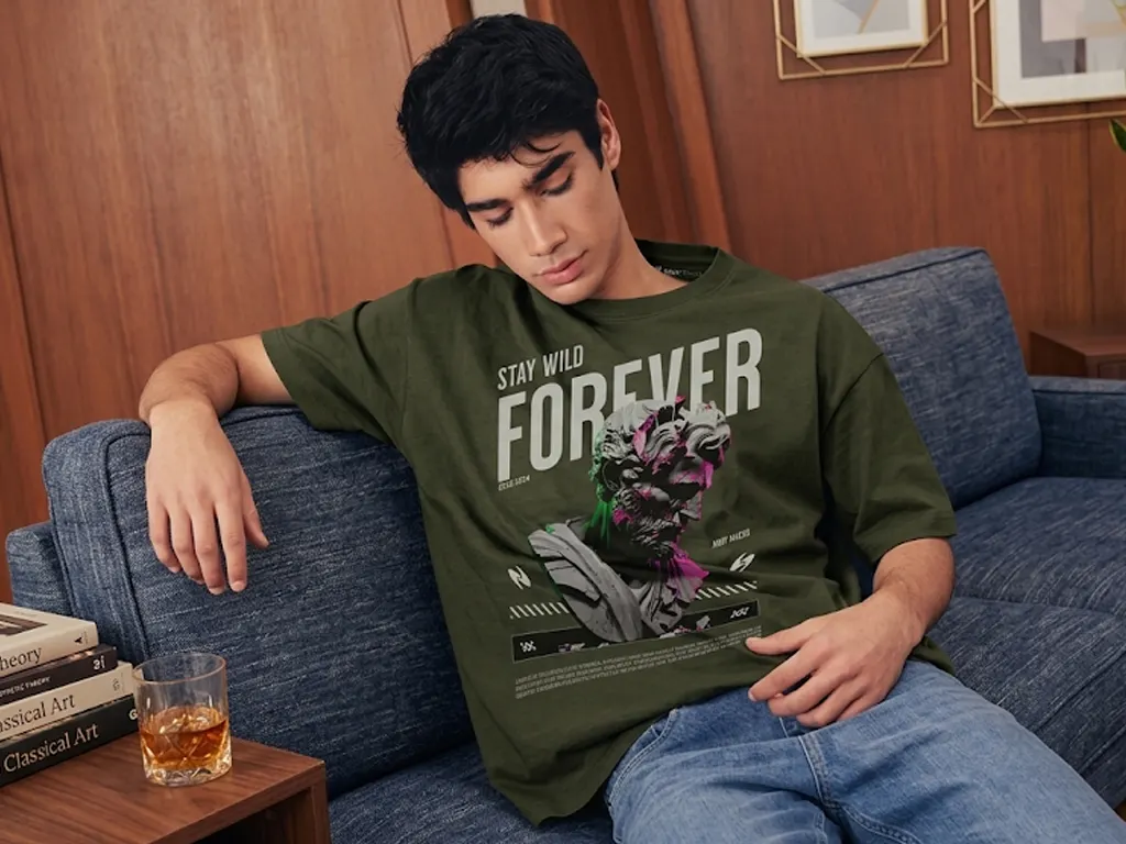 A young man wearing an olive green "Stay Wild Forever" graphic t-shirt relaxes on a blue sofa in a modern room with wood-paneled walls.