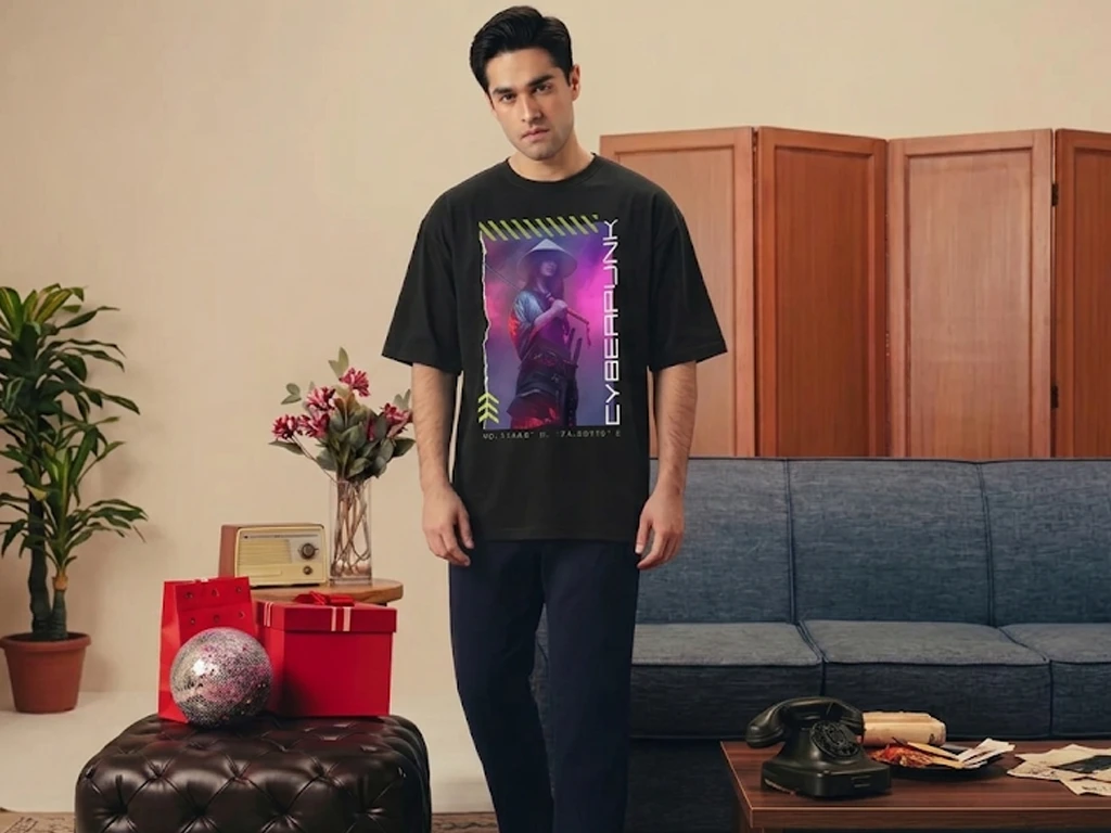 A man stands in a retro-modern living room wearing an oversized black t-shirt featuring a neon cyberpunk graphic of a character with a traditional hat and sword. The wide shot displays a blue sofa, a vintage rotary phone on a coffee table, and indoor plants.