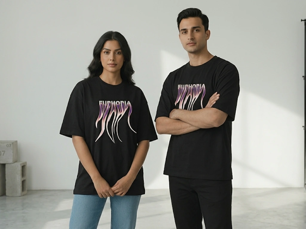 A full-body photograph of two models, a man and a woman, standing side-by-side in a minimalist studio. Both are wearing matching black, oversized, short-sleeve T-shirts with a stylized neon purple and white graphic print that resembles the word "EUPHORIA" in a distorted, wavy font on the front. The woman is on the left, looking directly at the camera with her hands clasped. She is wearing light-blue jeans and white sneakers. The man is on the right, also looking at the camera, with his arms crossed. He is wearing black jeans and black shoes. The background is simple and white, with a minimalist concrete block on the left and a large, empty, black-framed mirror or window on the right, partially visible. The studio floor is concrete.