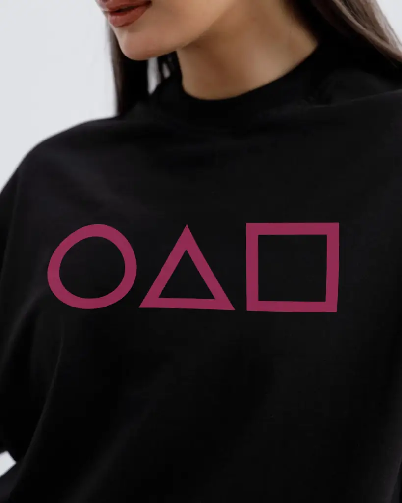 A close-up shot of a person wearing a black t-shirt featuring the iconic pink circle, triangle, and square symbols from Squid Game.
