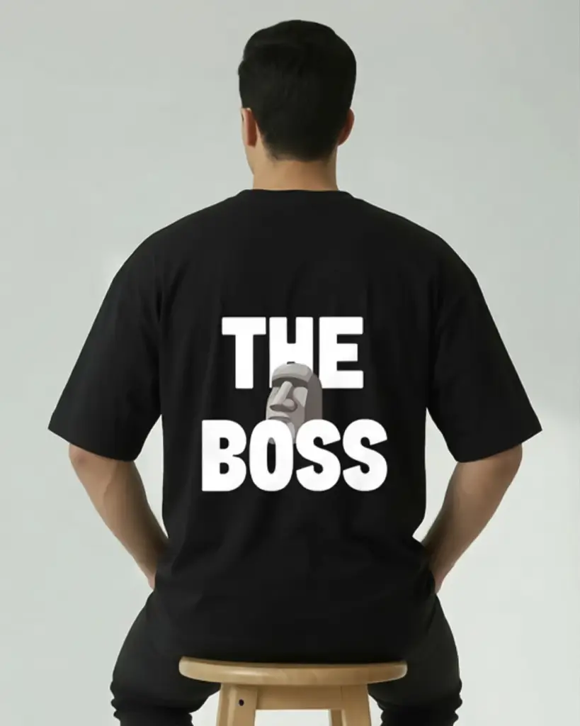 A view from behind of a man sitting on a round wooden stool against a plain light gray background. He is wearing an oversized black streetwear T-shirt that features bold white block text reading "THE BOSS" across the back. An illustration of a gray Moai stone statue head is integrated between the words, partially overlapping the text. His hands are resting on his upper thighs, and his dark hair is neatly styled.