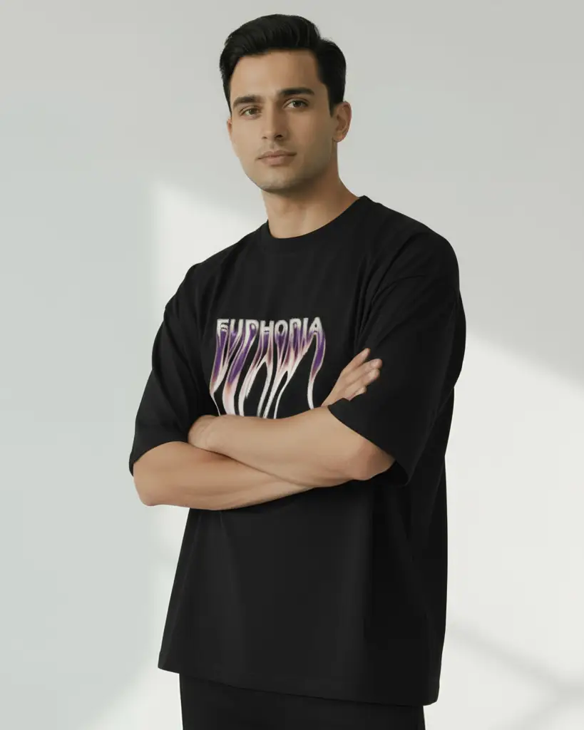 A man stands with his arms crossed wearing an oversized black t-shirt featuring a metallic, liquid-style graphic that reads "EUPHORIA" in dripping chrome and purple tones against a bright studio backdrop.