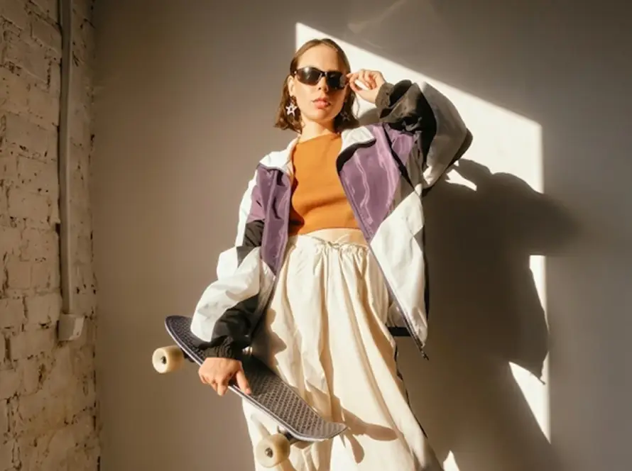 A wide landscape shot of a woman posing in a purple, black, and white color-blocked windbreaker, an orange crop top, and baggy white track pants while holding a black penny skateboard in a sunlit loft.