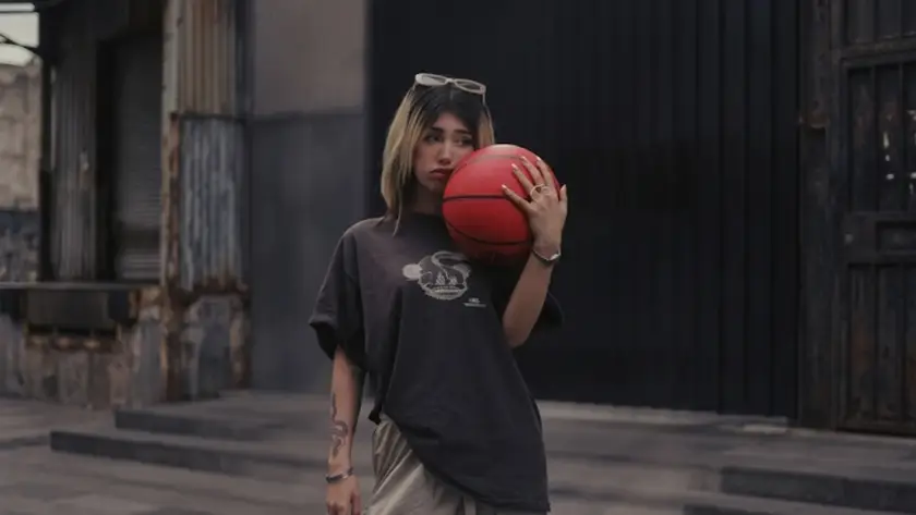 A young woman with blonde-highlighted hair wearing an oversized brown t-shirt and cargo pants, holding a red basketball against her shoulder in an urban setting.