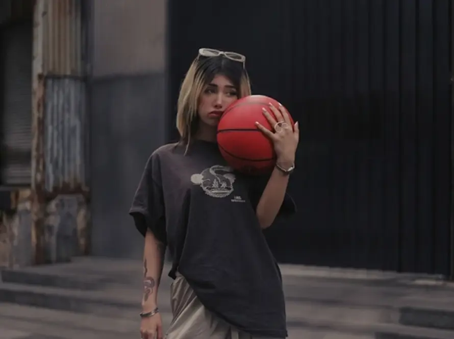 A young woman with blonde-highlighted hair wearing an oversized brown t-shirt and cargo pants, holding a red basketball against her shoulder in an urban setting.