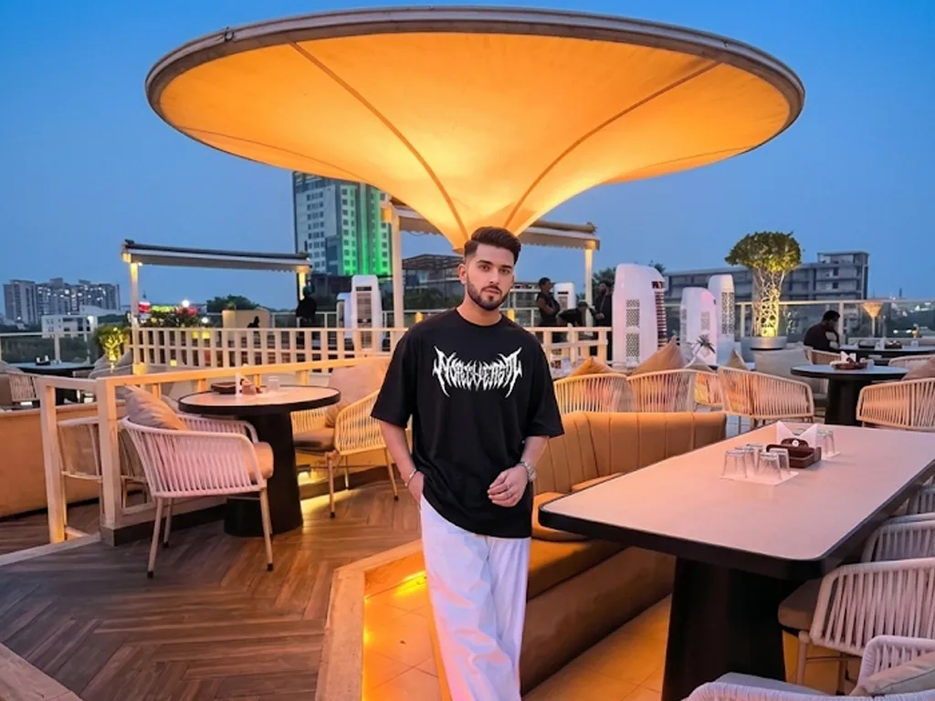 A wide landscape shot of a man standing on an illuminated rooftop cafe terrace at twilight, wearing an oversized black t-shirt with a spiky white metal-style graphic on the chest and wide-leg white trousers.