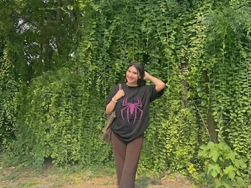 A wide landscape shot of a woman posing with one hand on her head in front of a massive wall of cascading green vines, wearing a black oversized t-shirt with a pink spider graphic and baggy brown trousers.