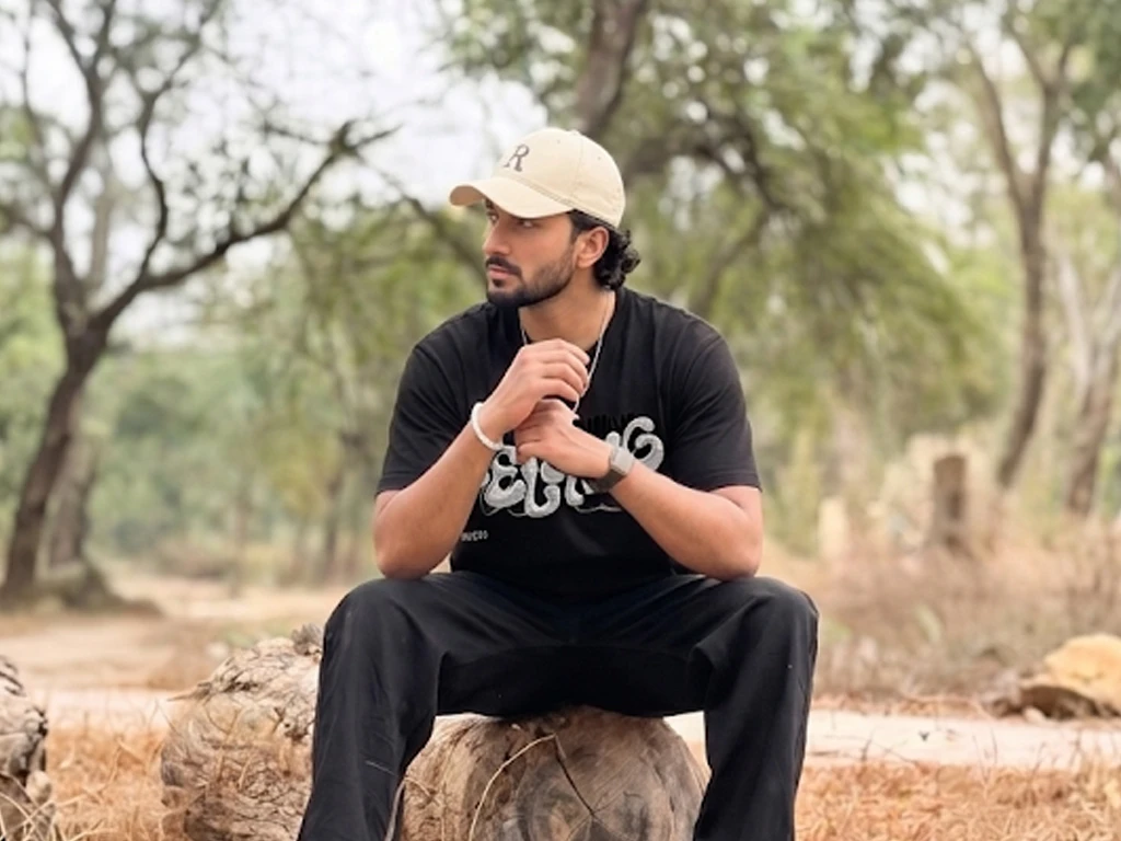 A man wearing a beige baseball cap and an oversized black t-shirt with a silver balloon-style graphic sits on a fallen log in a wide, scenic outdoor forest setting.