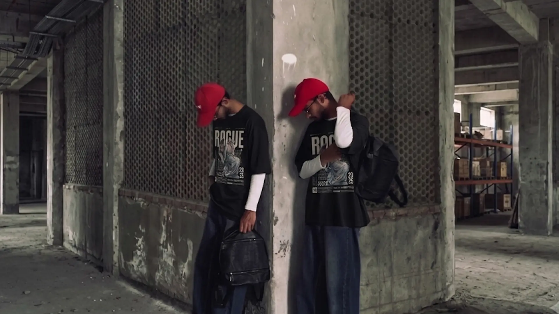 A wide landscape shot of two clones or individuals in identical street outfits leaning against opposite sides of a concrete pillar in an abandoned industrial warehouse, wearing oversized black "Rogue" t-shirts layered over white long-sleeve shirts, baggy jeans, and red caps.