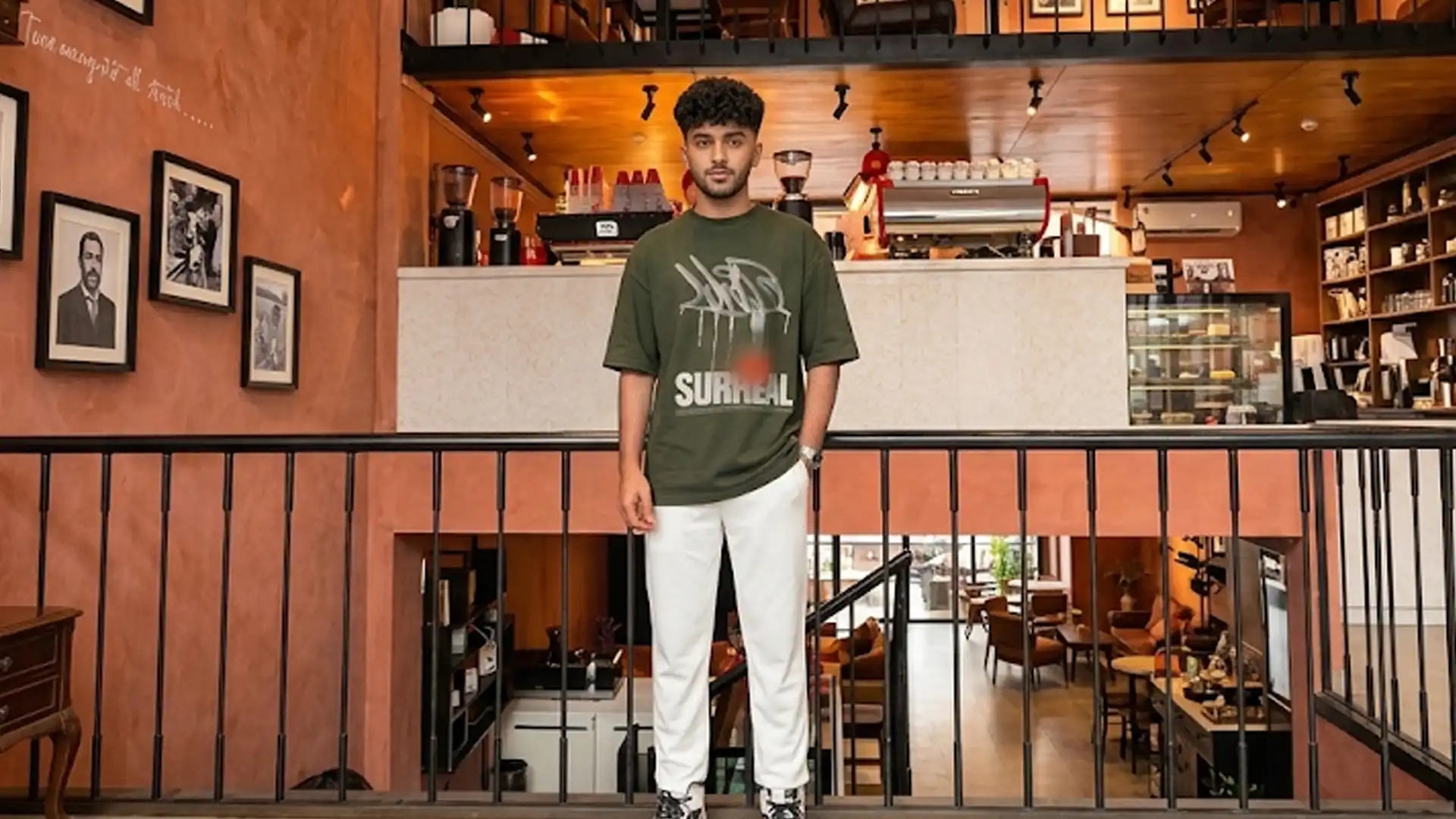 A wide landscape shot of a man standing behind a black metal railing on the middle level of a multi-tiered, terracotta-walled cafe, wearing an oversized olive green graphic t-shirt and white trousers.
