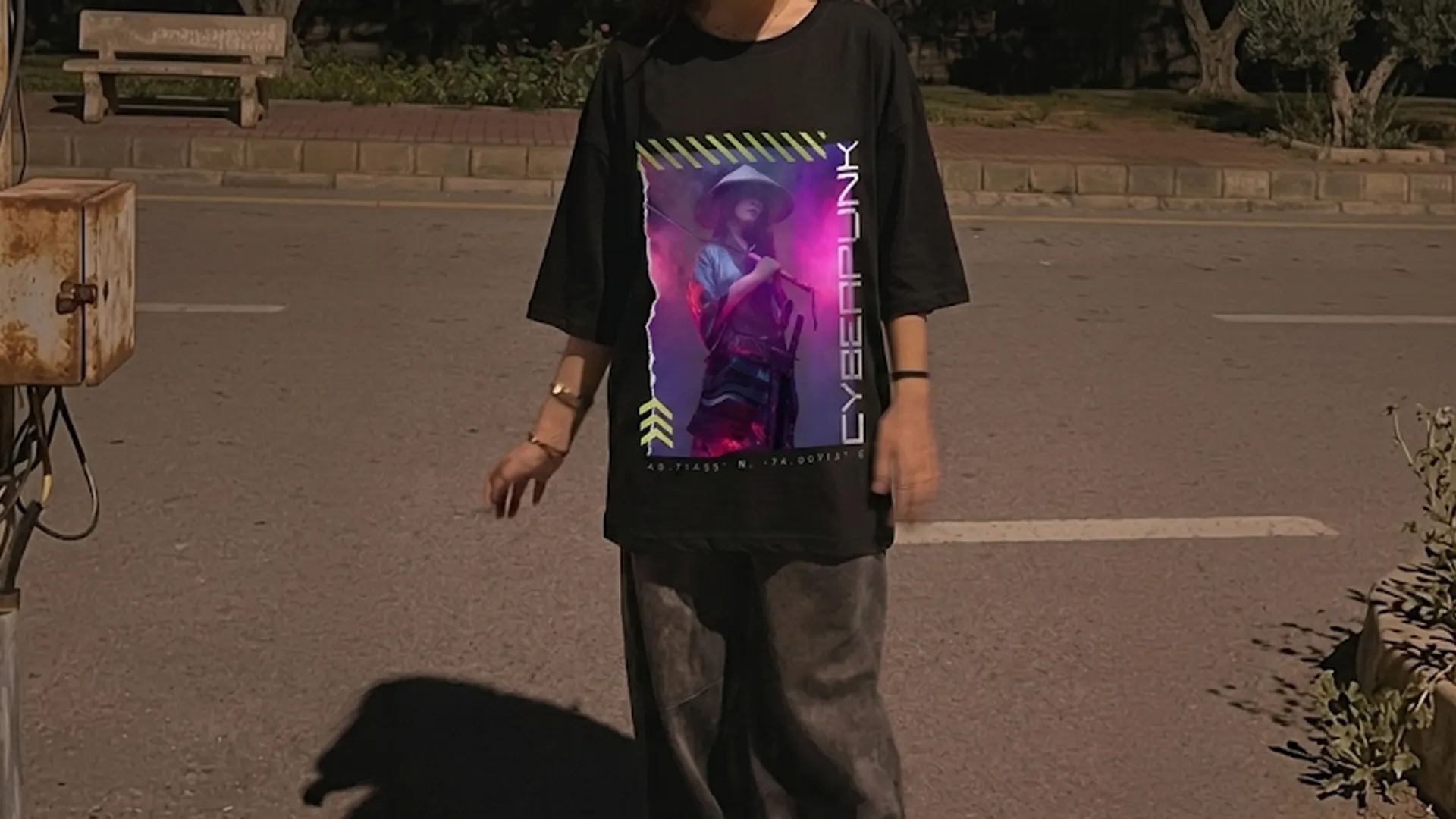 A wide landscape shot of a person standing on an asphalt road next to a weathered utility pole at dusk, wearing an oversized black t-shirt with a neon pink and purple cyberpunk graphic and baggy dark grey trousers.