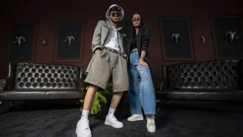 A powerful, low-angle photograph of two young, stylish models standing confidently in an edgy, luxurious gothic room. The man on the left wears a bucket hat, sunglasses, a grey coat over a white tee, and wide-leg shorts. The woman on the right wears red sunglasses, a black blazer, a black top, and ripped wide-leg jeans. Both models wear white sneakers and pose with their hands on their hips or in their pockets. They are standing on a dark carpet between two black leather tufted Chesterfield sofas. Behind them, a deep burgundy wall is decorated with multiple framed, stylized dark ram skulls.
